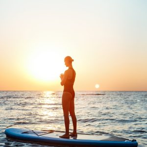 Silhouette of young beautiful girl practicing yoga on surfboard in sea at sunrise.